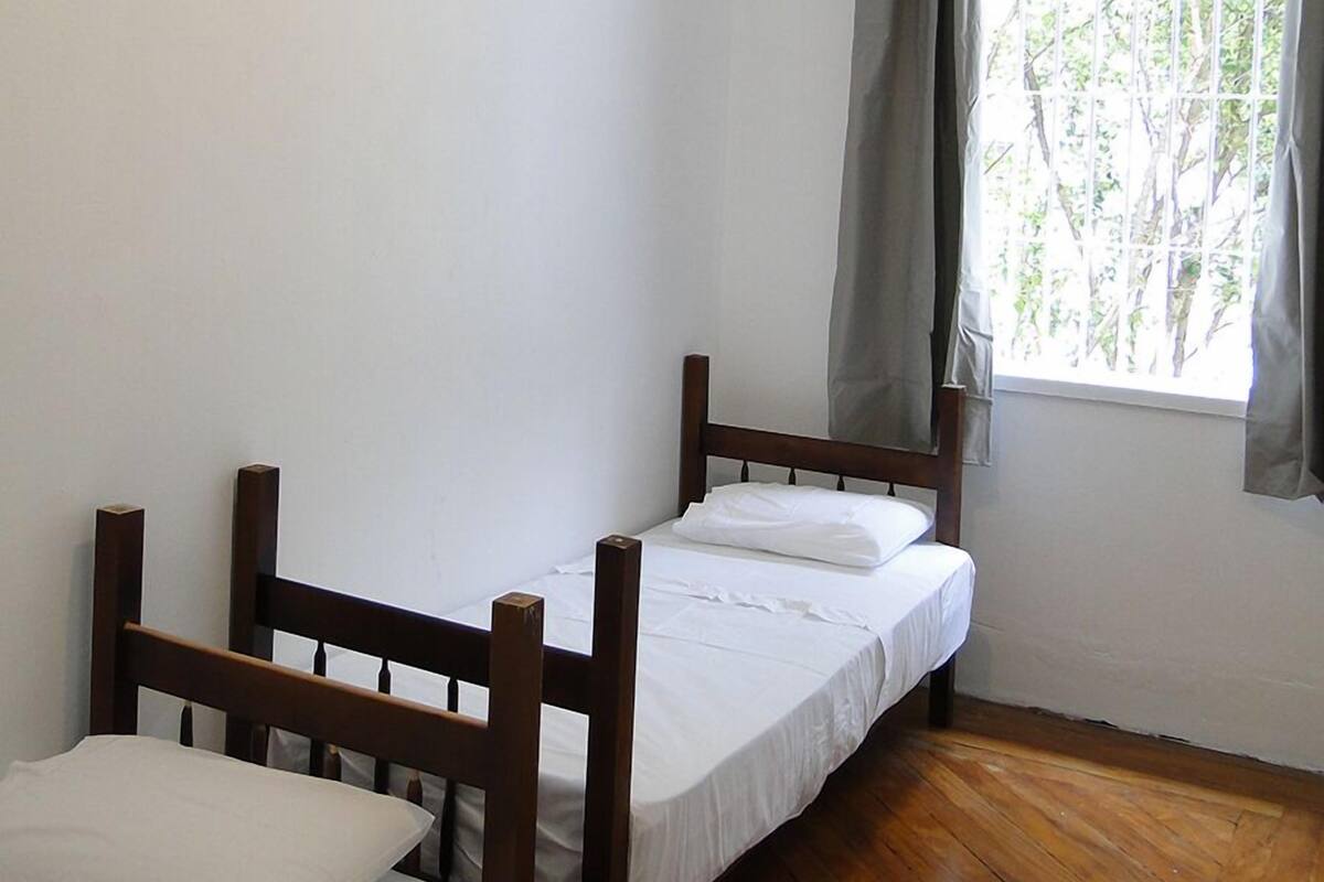 Eine kleine, einfache Unterkunft mit zwei Holzbetten, einem Fenster mit Vorhängen und einem Deckenventilator.