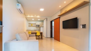 A modern living room with a sofa, a flat-screen TV mounted on the wall, and a kitchen in the background. - Hotel Brasil (Penha)