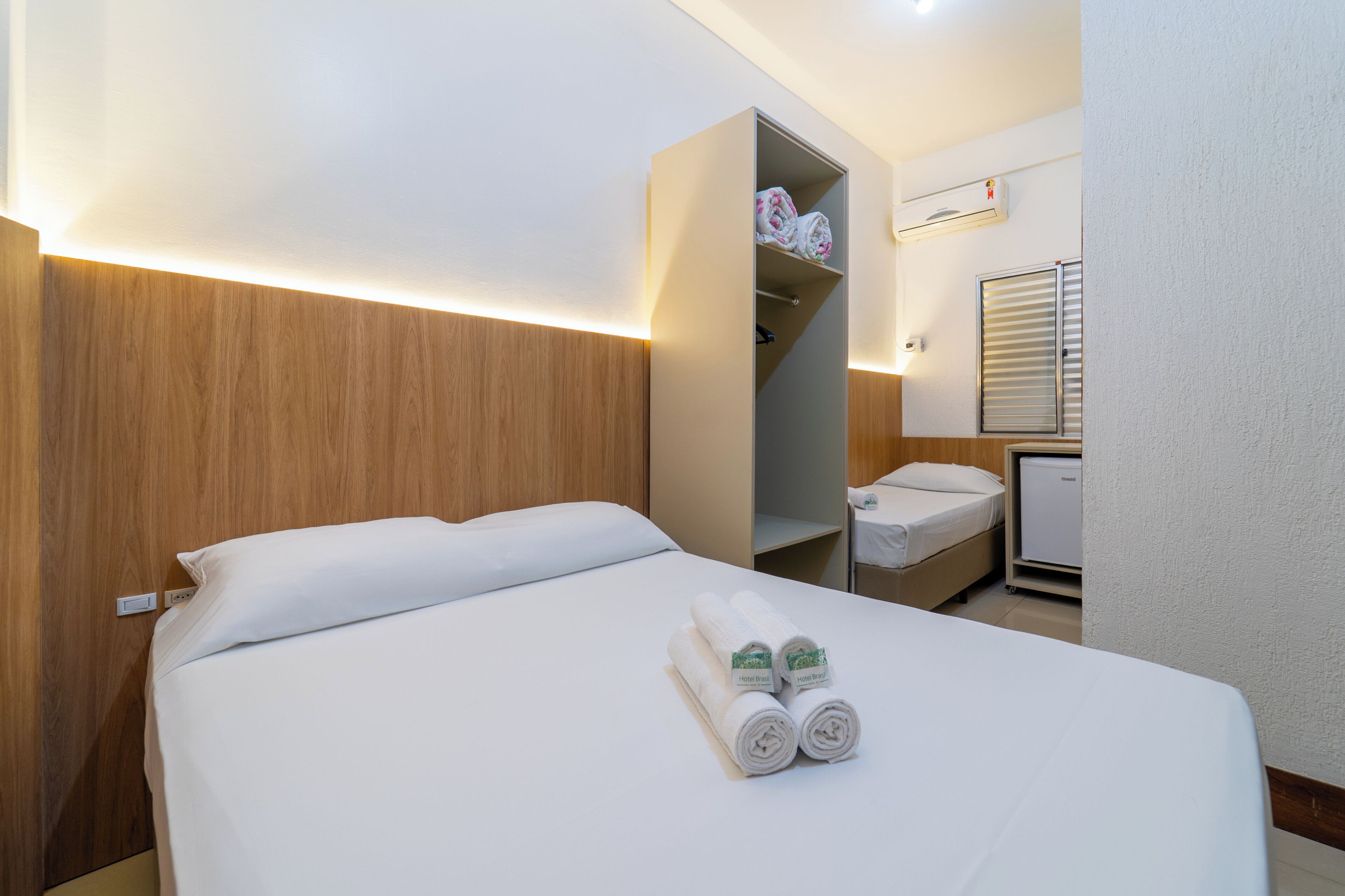 A hotel room with a bed, a wardrobe, a small desk with a chair, and a window with blinds.
