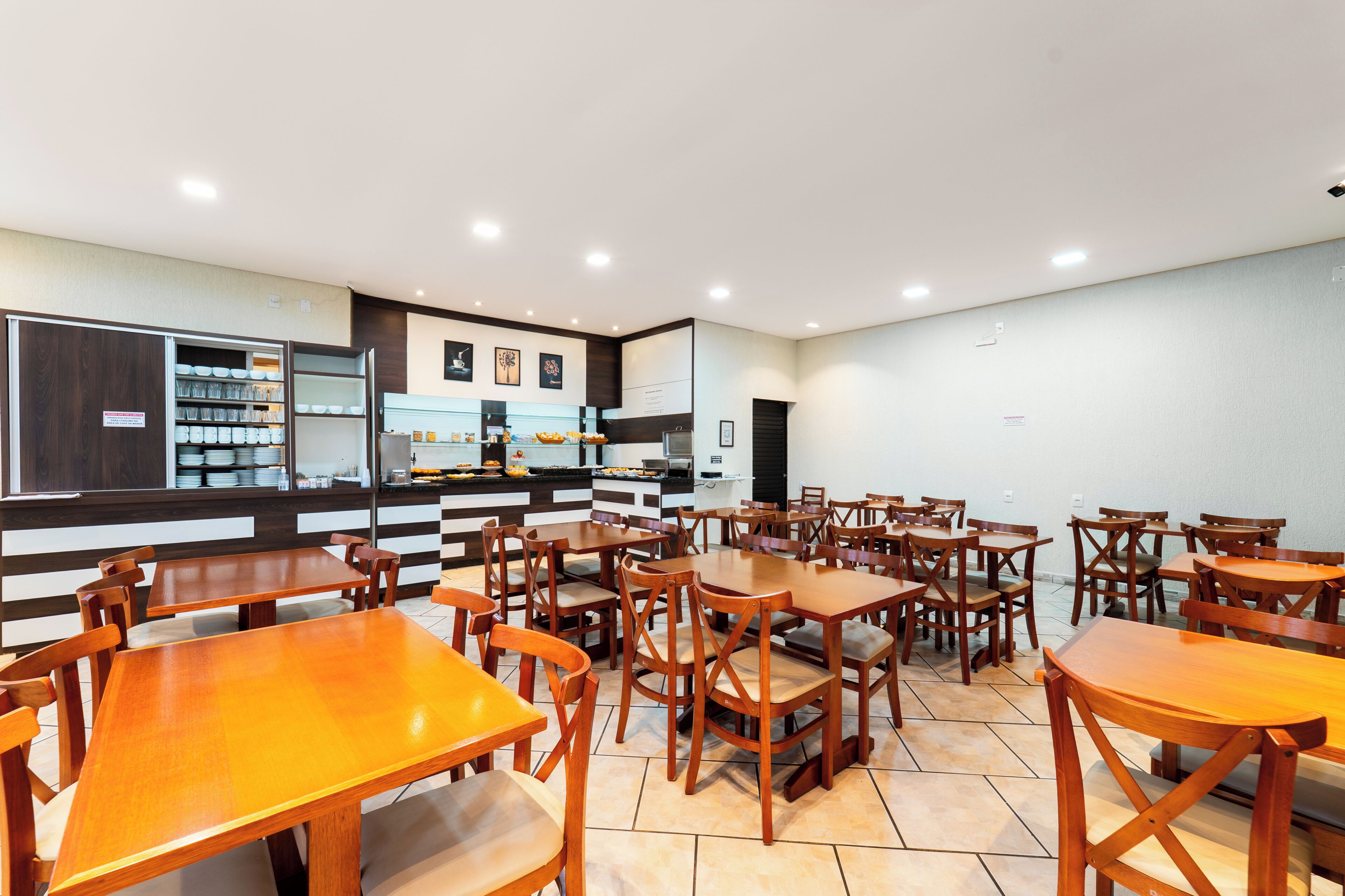 A restaurant with multiple wooden tables and chairs, a counter with food items, and a wall with framed pictures.