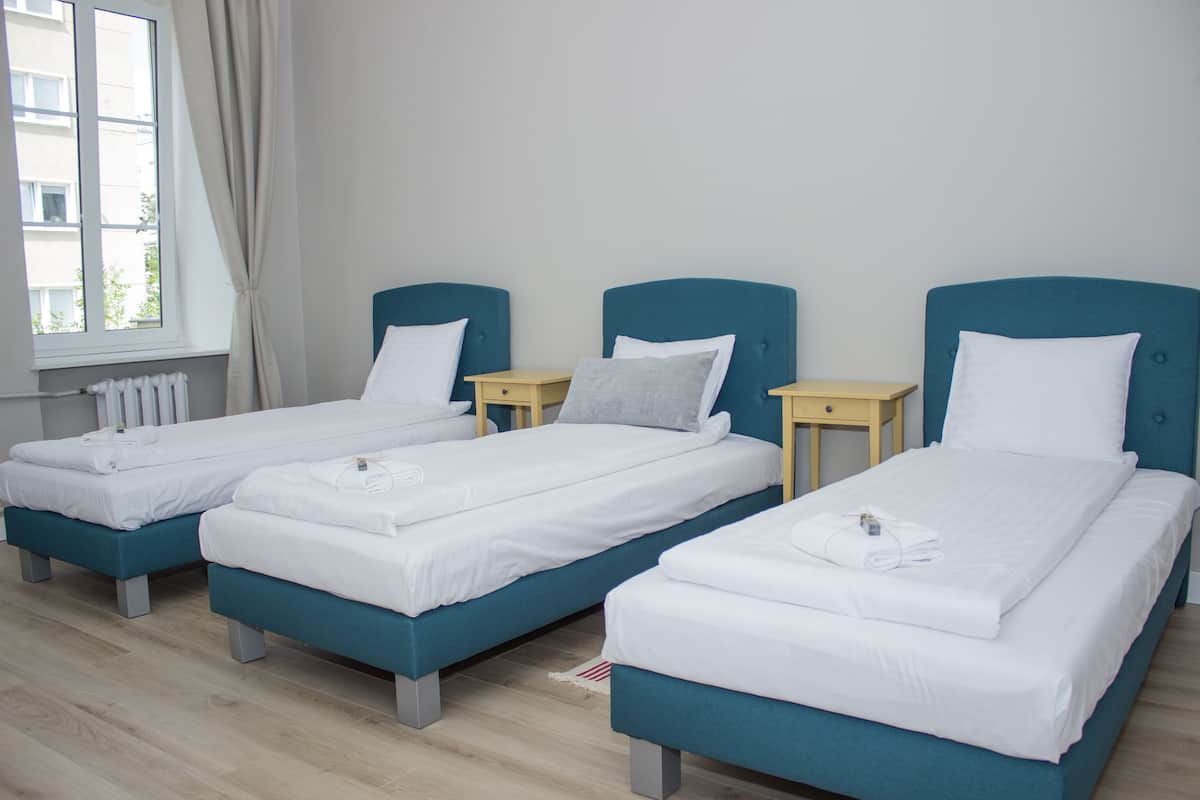 Une chambre d’hôtel avec trois lits simples, chacun doté d’une tête de lit bleue et de draps blancs. Il y a une fenêtre avec des rideaux sur le côté gauche.