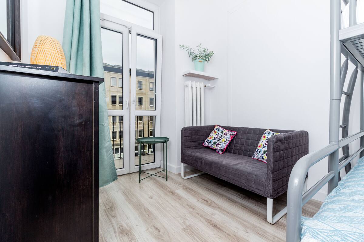 Une pièce moderne avec un bureau en bois foncé, un canapé gris agrémenté de coussins colorés, un petit tabouret vert et une plante en pot posée sur une étagère blanche.