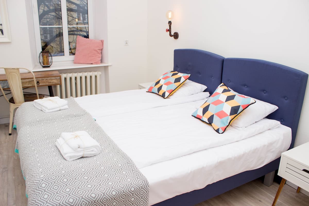 Un lit double avec une tête de lit bleue, deux coussins colorés, une couette blanche et un couvre-lit gris à motifs.