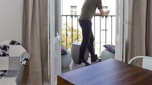 Un hombre está de pie junto a una puerta de balcón, una mesa de madera y una silla con estructura roja.