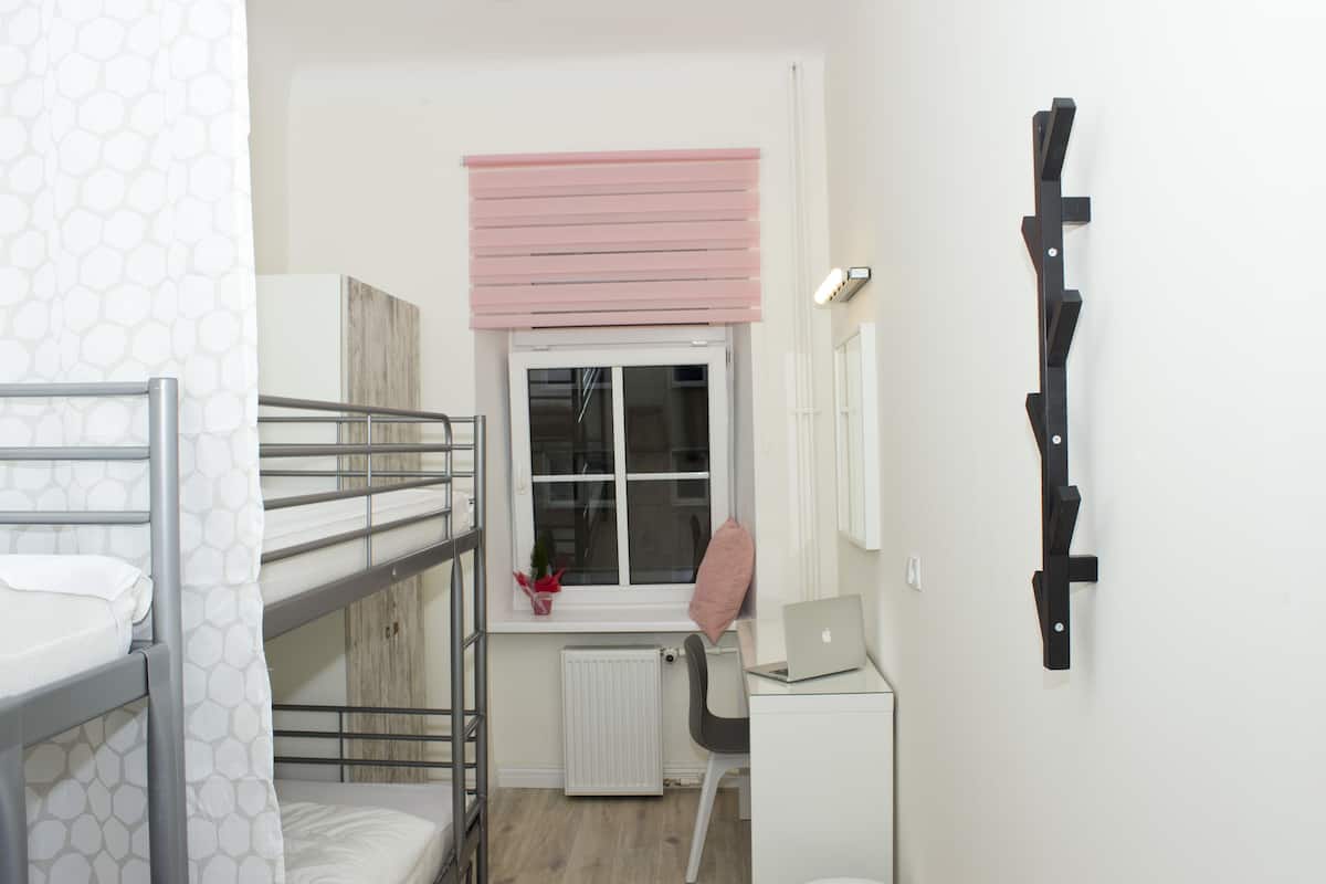 Une chambre de dortoir avec des lits superposés, un bureau avec un ordinateur portable, un radiateur, une fenêtre avec des stores et un porte-manteau fixé au mur.