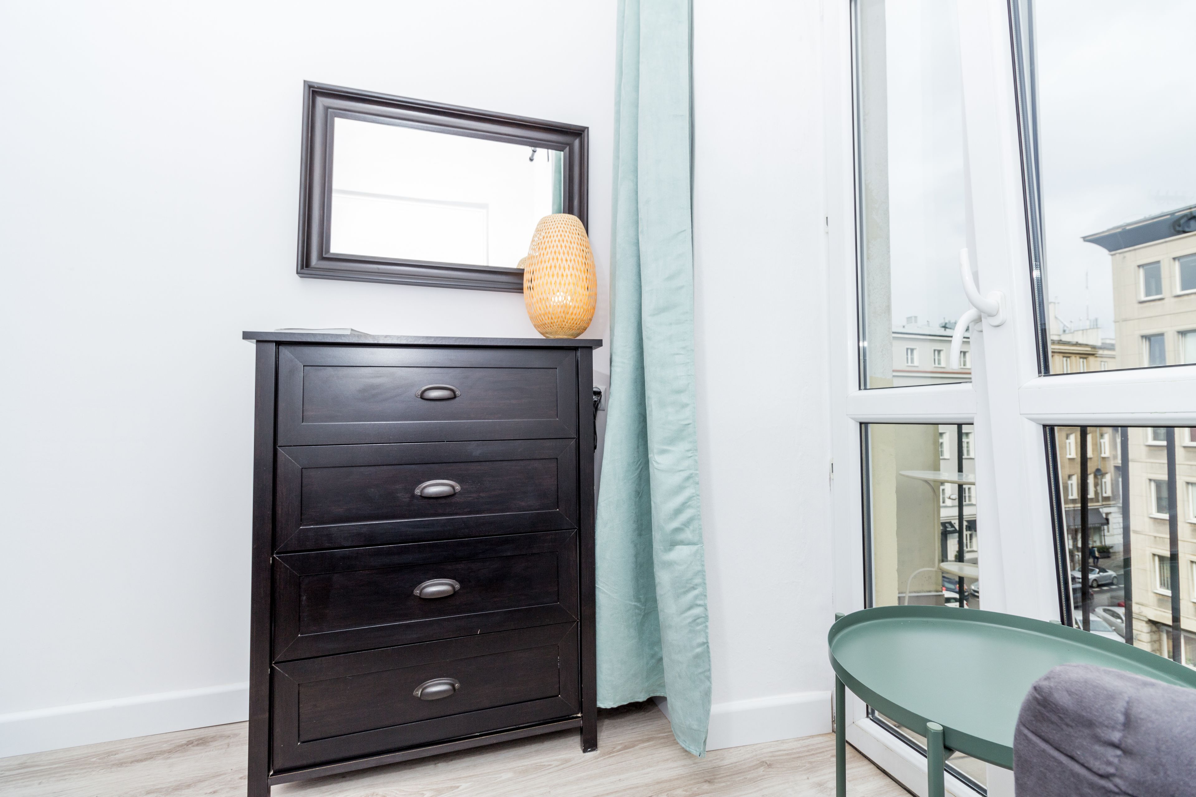 Une pièce avec une commode en bois foncé, un fauteuil vert, une fenêtre donnant sur des immeubles et un rideau vert clair.