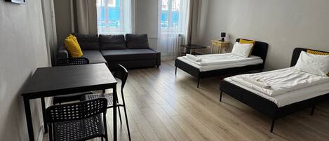 A room with a sofa, two single beds, a table with chairs, and a small table with a lamp.