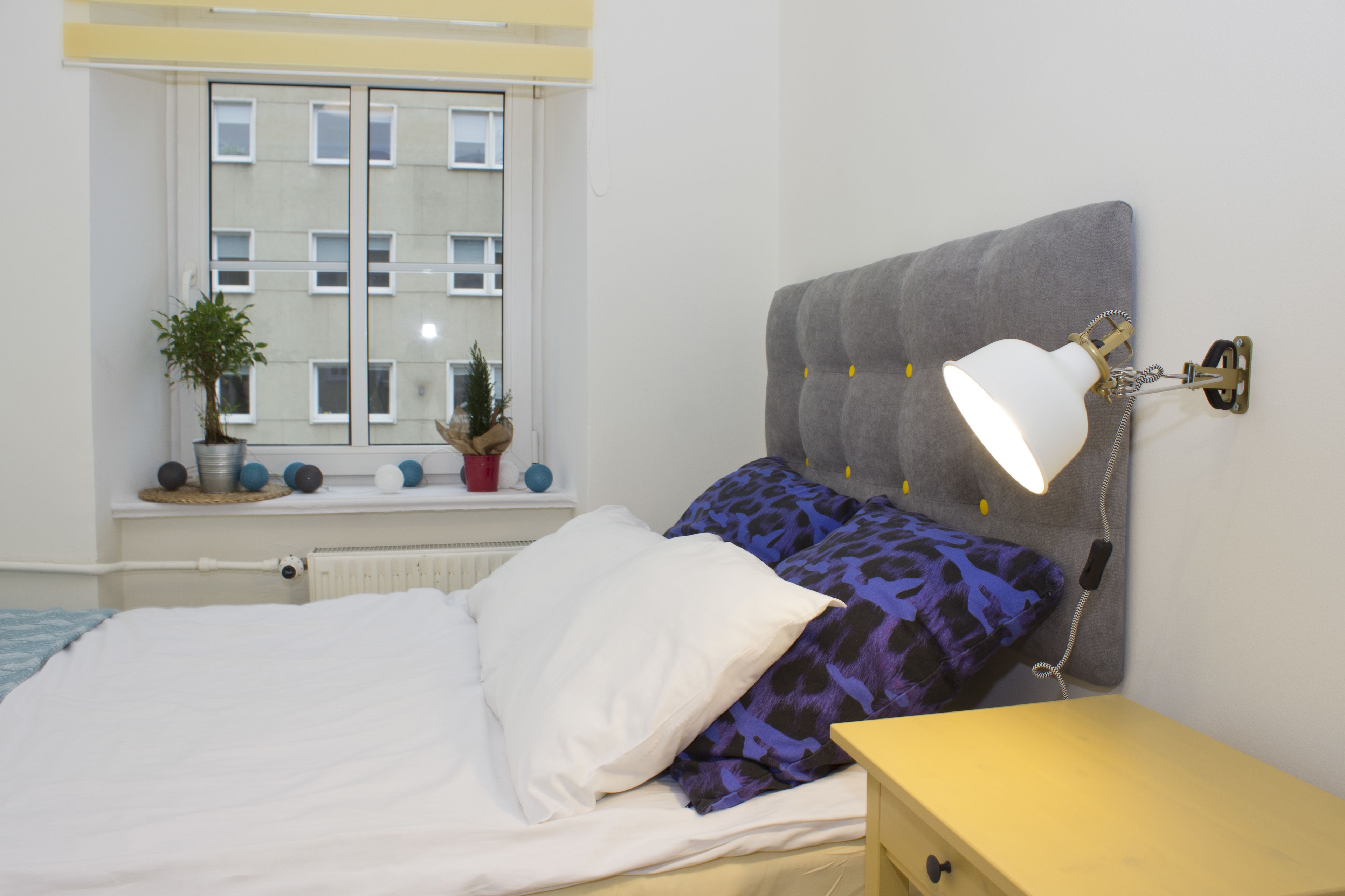 Un dormitorio con cama, una mesita de noche, una lámpara, una ventana con vistas a edificios y una planta en maceta en el alféizar.