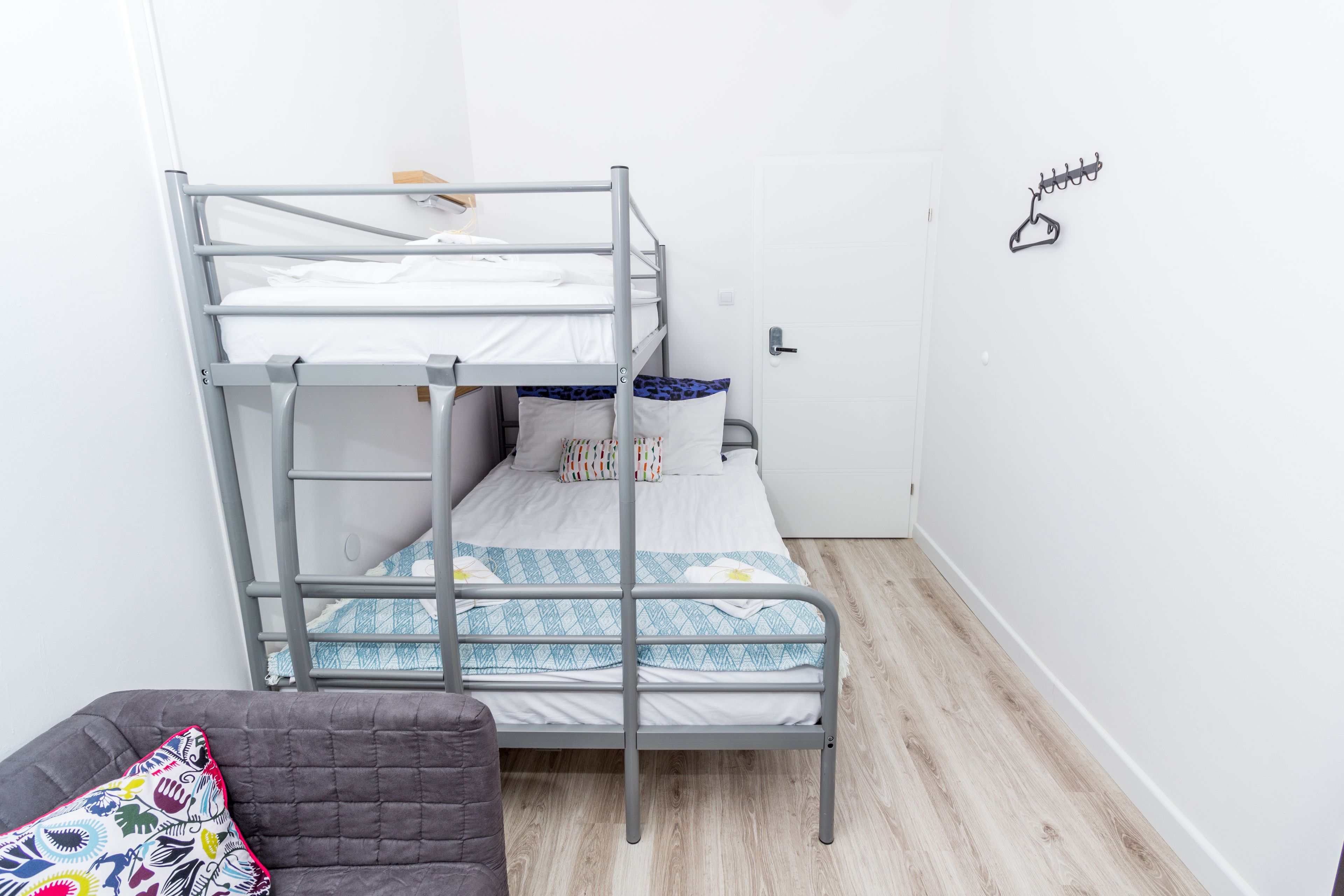 A bunk bed with a lower bunk, a grey couch with a colorful pillow, and a door with a coat hanger.
