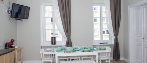 Un coin repas avec une table blanche dressée pour six personnes, un téléviseur fixé au mur et une fenêtre avec des rideaux.