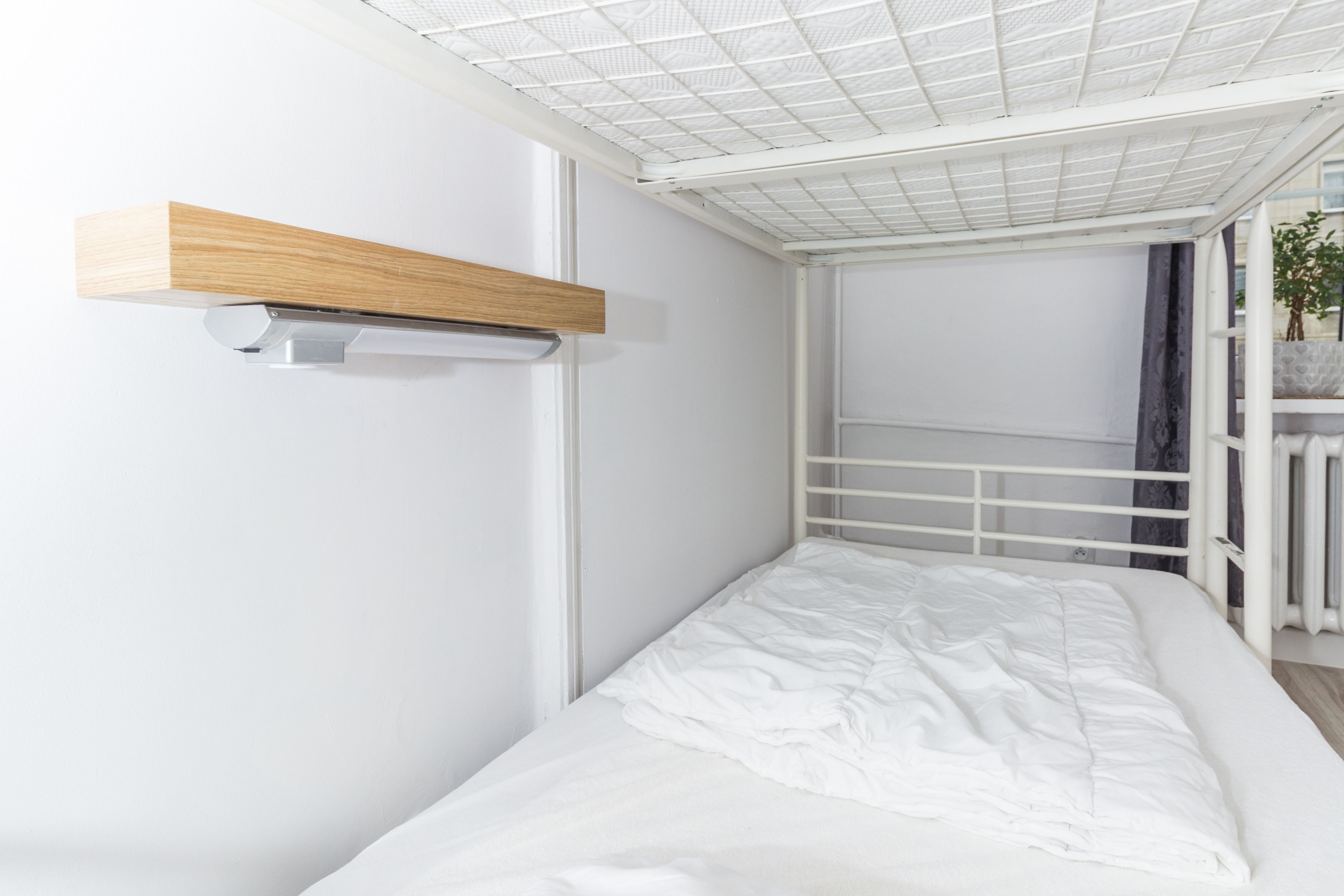 A small, white, single-bed room with a wooden shelf above the bed and a white railing on the window.