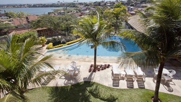 A pool with lounge chairs and palm trees, surrounded by a well-maintained lawn and a view of a body of water and distant buildings. - Bon Bini Pousada (Búzios)