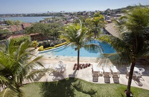 Una piscina con sillones de descanso y palmeras, rodeada de un césped cuidado y con vista a una masa de agua y edificios al fondo.