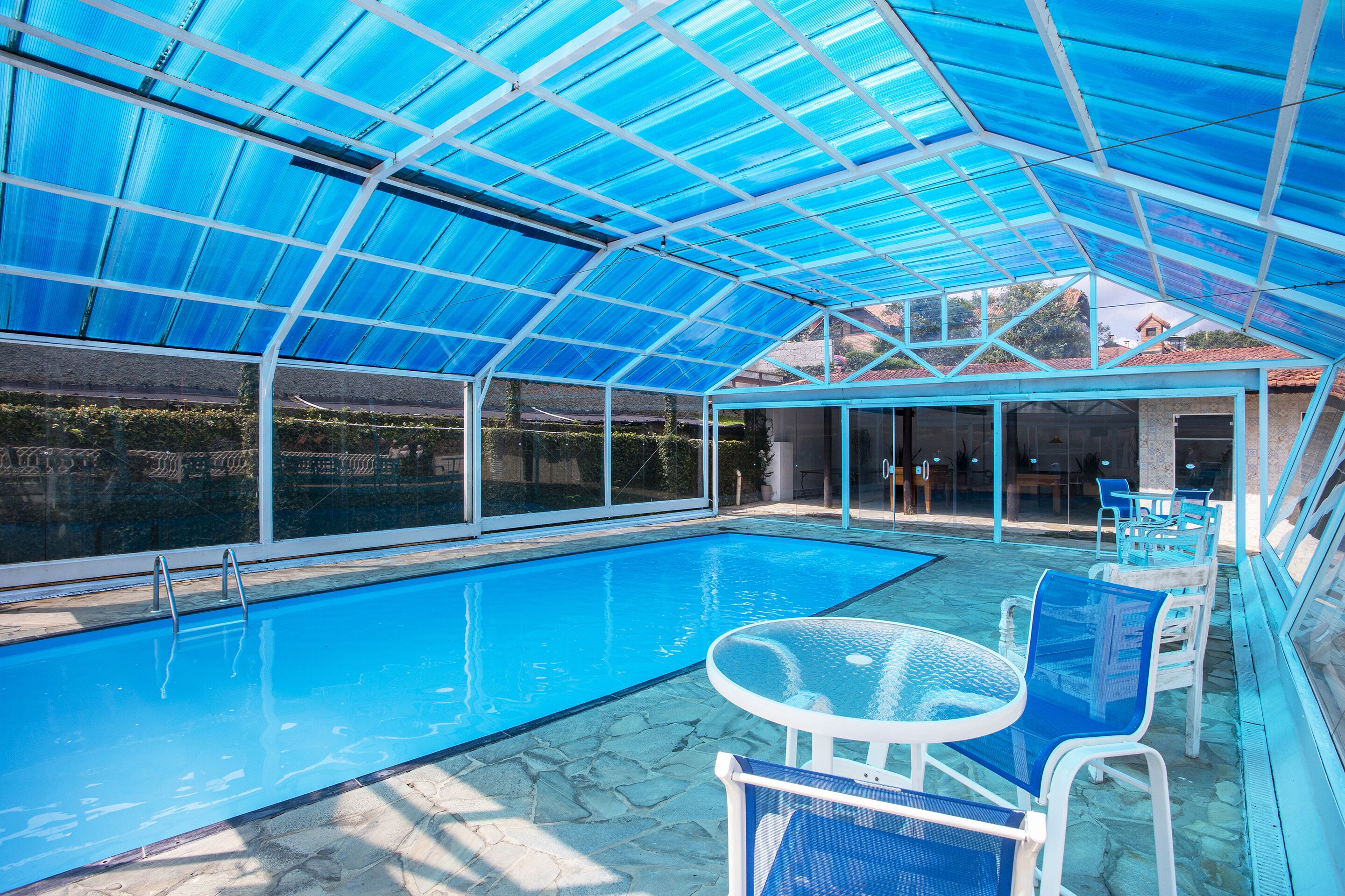 Ein überdachter Swimmingpool mit Glaswänden und einem blauen Dach, umgeben von Steinböden und blauen Stühlen.