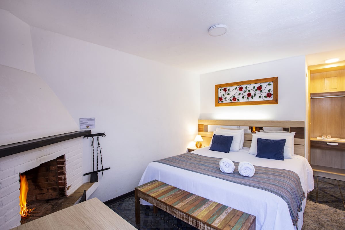 Una habitación de hotel con chimenea, una cama con manta a rayas, dos almohadas, una mesita de noche y una obra de arte colgada en la pared.