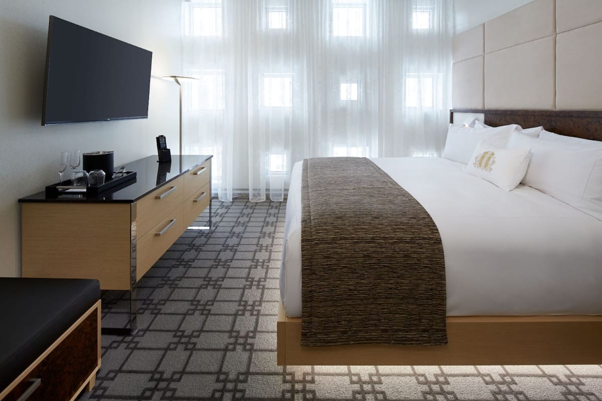 Habitación de hotel con cama, televisión, escritorio con cajones, lámpara y alfombra estampada.