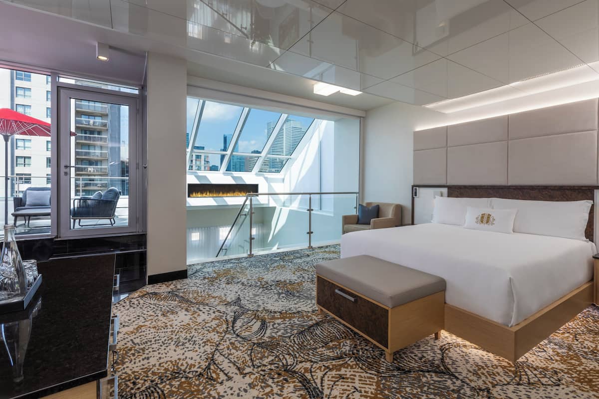 Una habitación de hotel moderna con una cama grande, un balcón con vistas a la ciudad, una zona de estar con una mesa y una alfombra estampada.