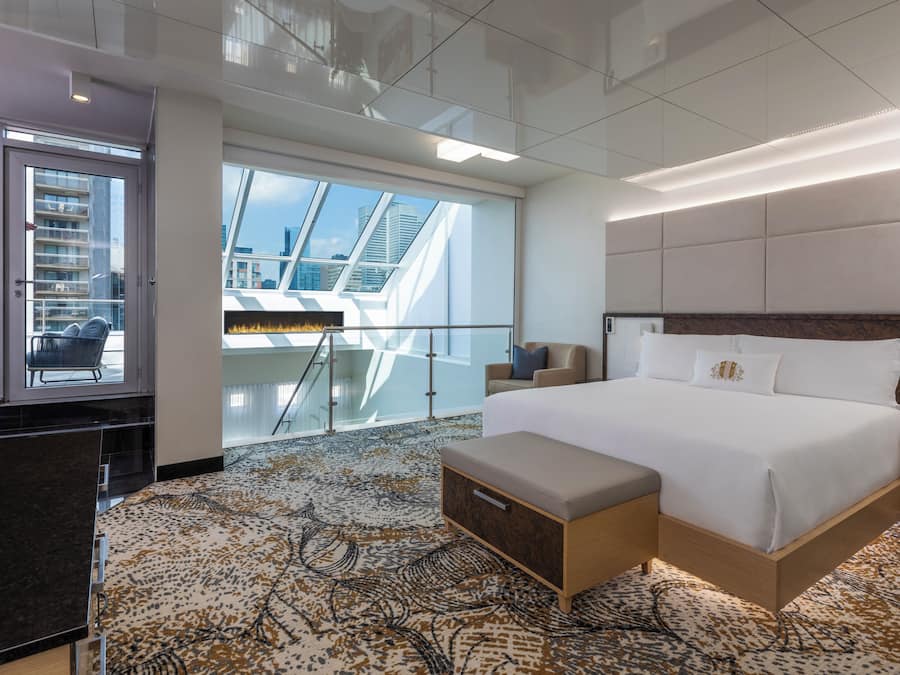 Una habitación de hotel moderna con una cama grande, un balcón con vistas a la ciudad, una zona de estar con una mesa y una alfombra estampada.