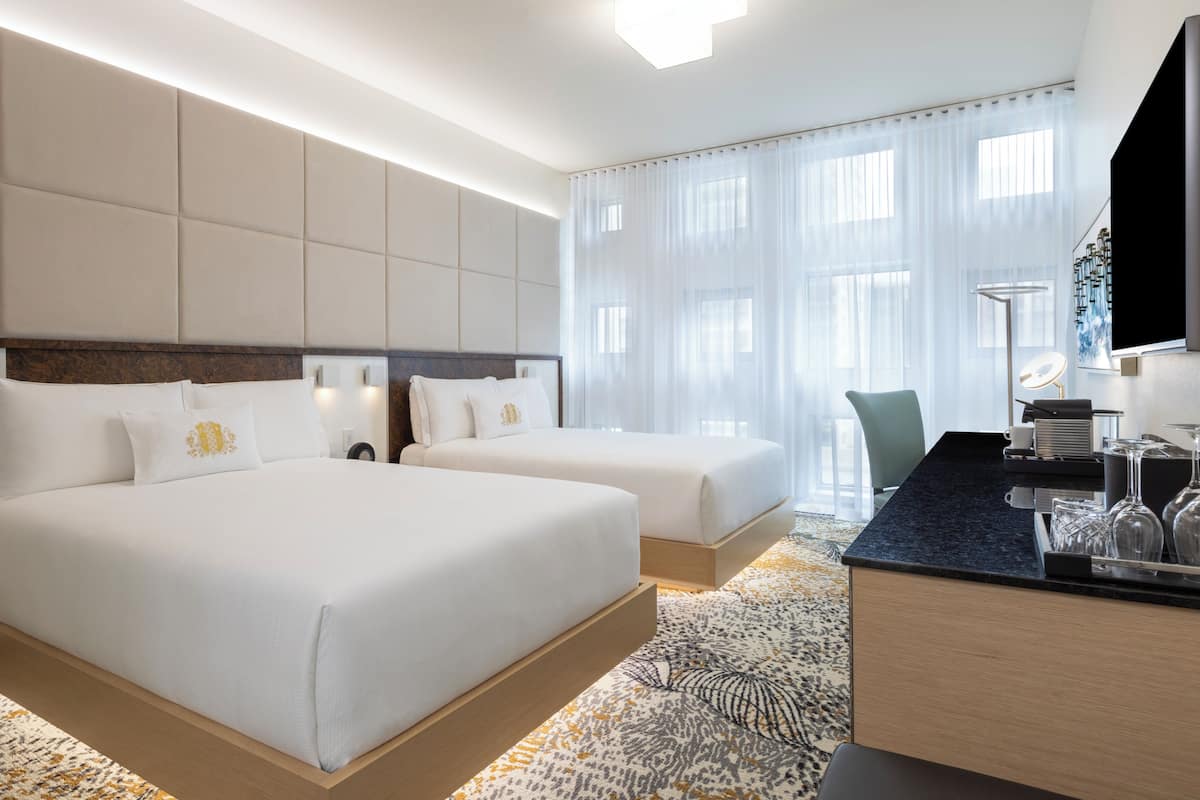 Una habitación de hotel con dos camas, un escritorio con lámpara y un ventanal con cortinas translúcidas.