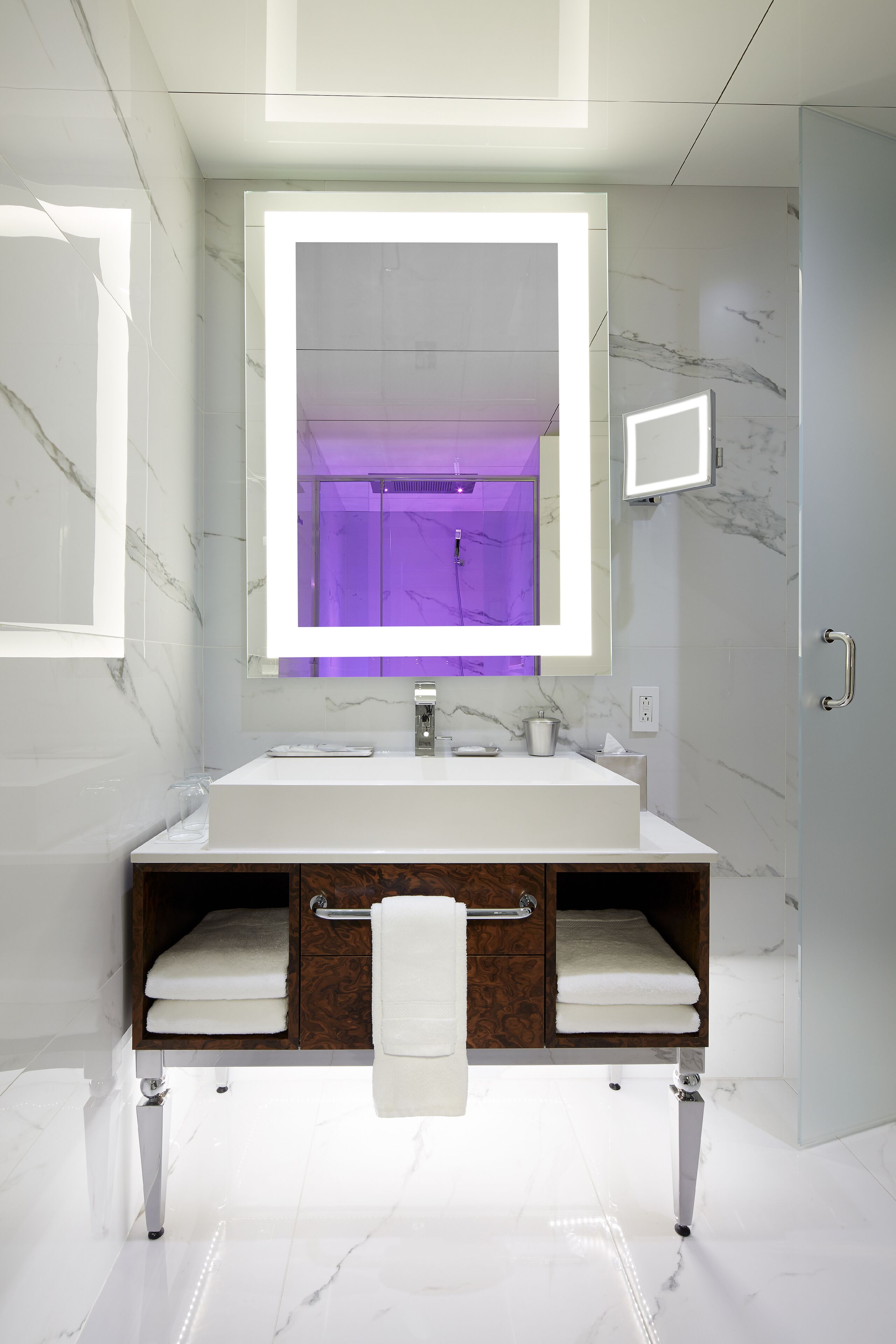 Un baño moderno con un lavamanos grande de color blanco, un espejo con fondo iluminado en morado y un mueble de tocador de madera con dos barras para toallas.