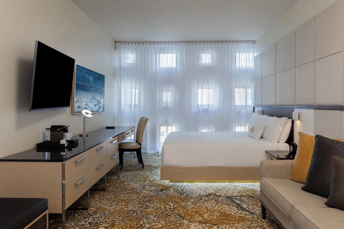 Habitación de hotel con cama, escritorio, silla, televisor y un ventanal con cortinas ligeras.