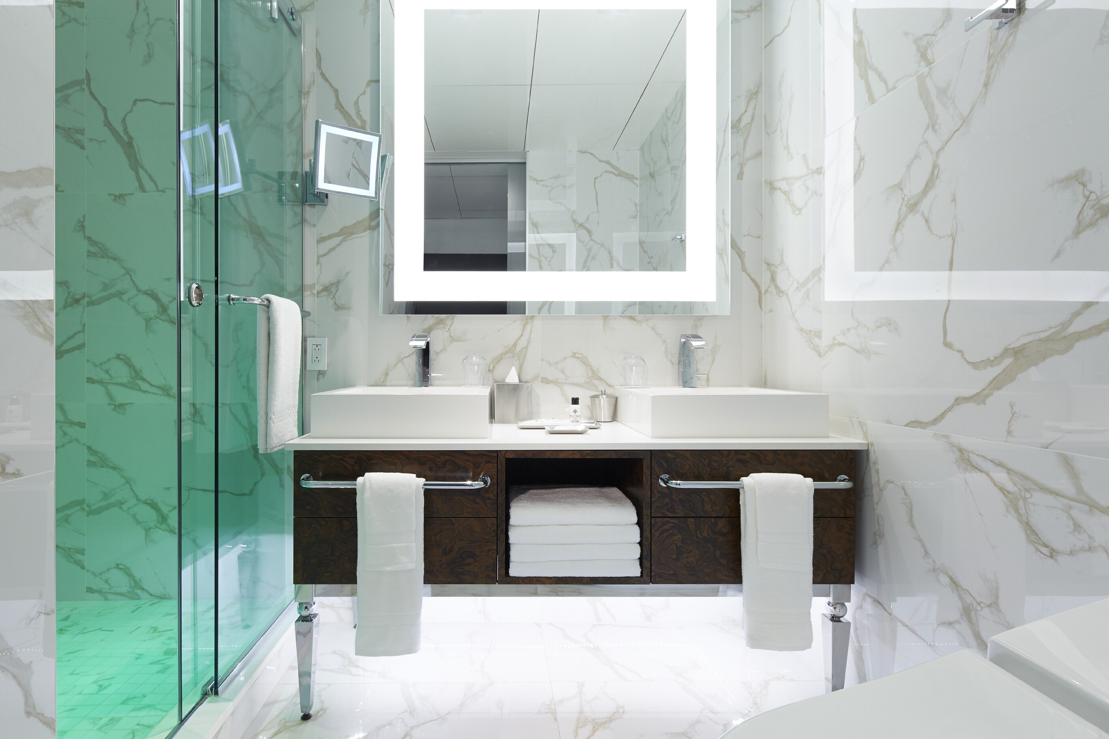 Un baño moderno con cabina de ducha de vidrio, pared con azulejos de mármol, espejo grande y tocador doble.