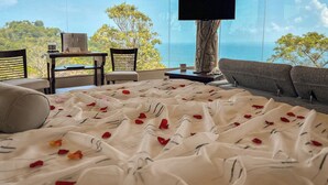 Ein Schlafzimmer mit einem großen Bett, einem Schreibtisch und einem Sessel, mit Blick auf eine schöne Landschaft aus Meer und Bäumen.
