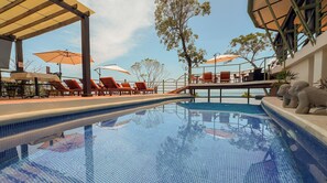 Ein Schwimmbecken mit Liegestühlen und Sonnenschirmen, umgeben von einer überdachten Fläche mit Blick auf den Ozean.
