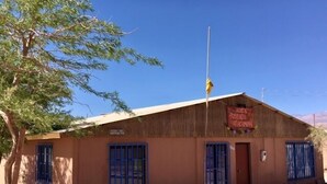 A single-story building with a thatched roof, blue metal bars on the windows, and a flagpole with a flag. - Hostal Possada Atacameña (San Pedro de Atacama)