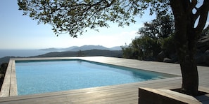 A rectangular swimming pool with a wooden deck, surrounded by trees and overlooking a mountainous landscape. - Résidence Bianca Casa (Porto-Vecchio)