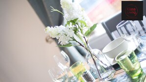 Une table à manger dressée avec de l’assiette blanche, des verres transparents et un vase contenant des fleurs blanches.