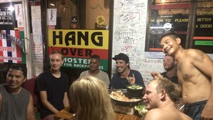 A group of people sitting around a table in a casual dining area with a sign that reads "HANG OVER HOSTEL FOR BACKPACKERS". - Hangover Hostel (Ko Phi Phi)