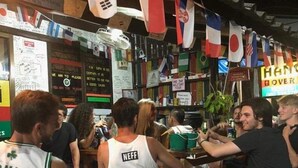 A group of people sitting at a bar with various flags hanging above. - Hangover Hostel (Ko Phi Phi)