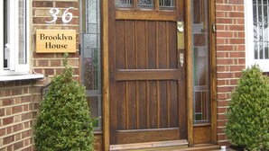 Een houten voordeur met een glasraam erboven, een bakstenen muur en een bordje met de tekst 'Brooklyn House 36'.