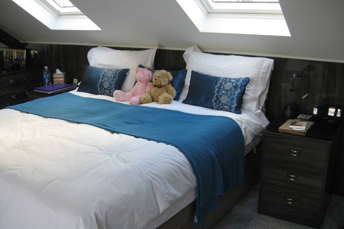 Ein Schlafzimmer mit einem großen Bett, zwei Kissen, einem Teddybären, einem Nachttisch und einem Oberlicht.