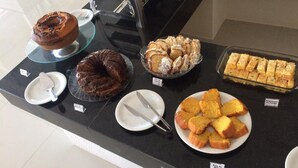 A buffet counter with various desserts including a chocolate cake, a bundt cake, a slice of cheesecake, and several types of bread. - Hotel Caju de Ouro (Feira de Santana)