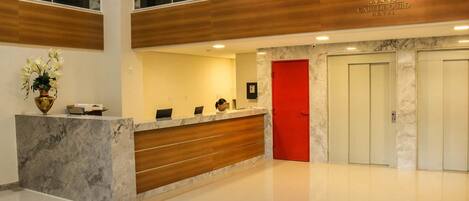 Zona de recepción de un hotel con mostrador de mármol, una recepcionista, una puerta roja y un letrero que dice "CAJU DE OURO HOTEL".