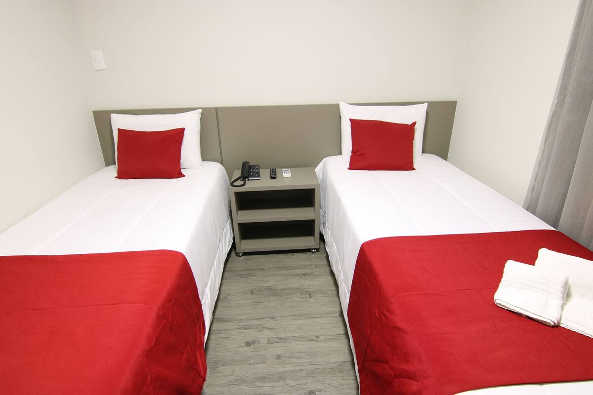 Ein Hotelzimmer mit zwei Einzelbetten, jeweils mit einem roten und weißen Kopfkissen, einem kleinen Nachttisch mit Telefon und einer roten Bettdecke.