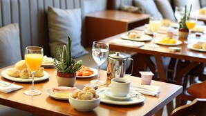 A breakfast setting with a variety of dishes, including pastries, fruits, and beverages, arranged on a wooden table in a modern dining area. - Lagoa Flat Hotel (Lagoa da Prata)