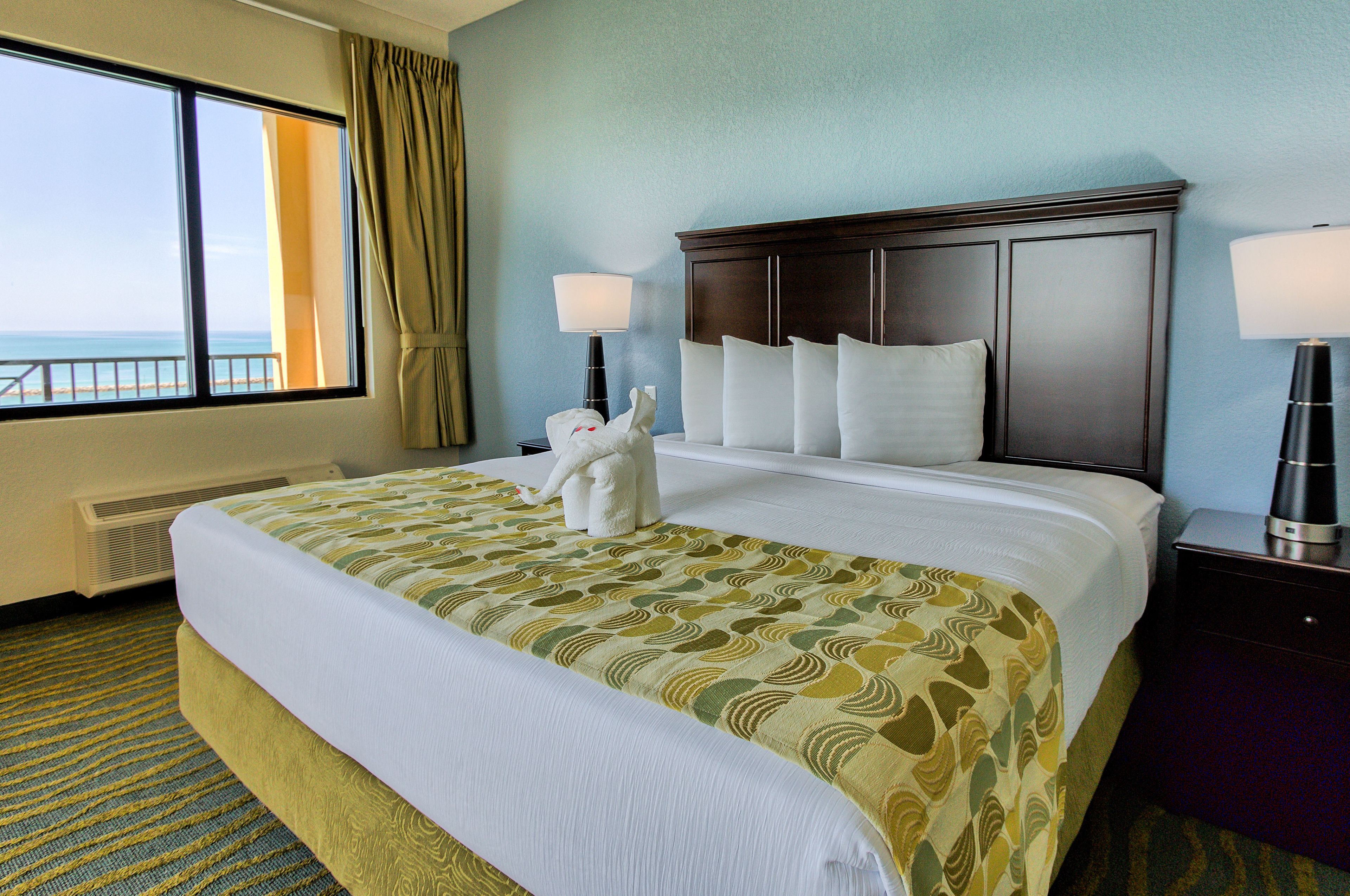 A hotel room with a large bed, two bedside lamps, a view of the ocean, and a window with curtains.