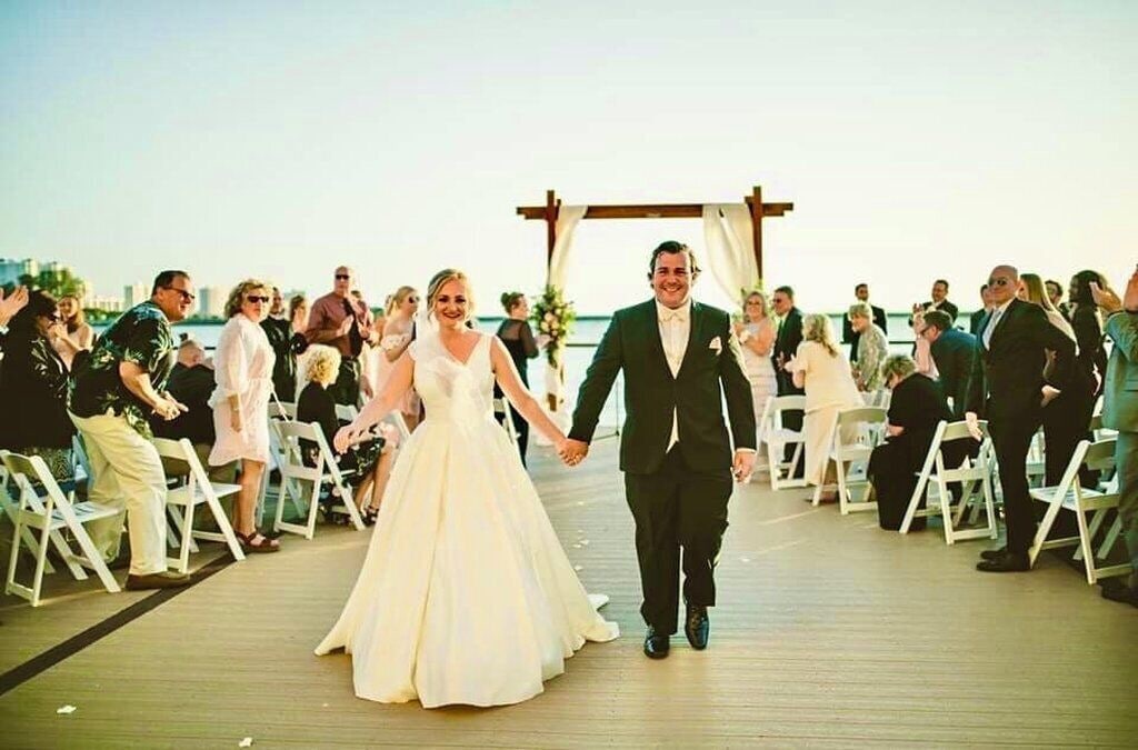 A wedding ceremony on a wooden deck with guests seated on white chairs.