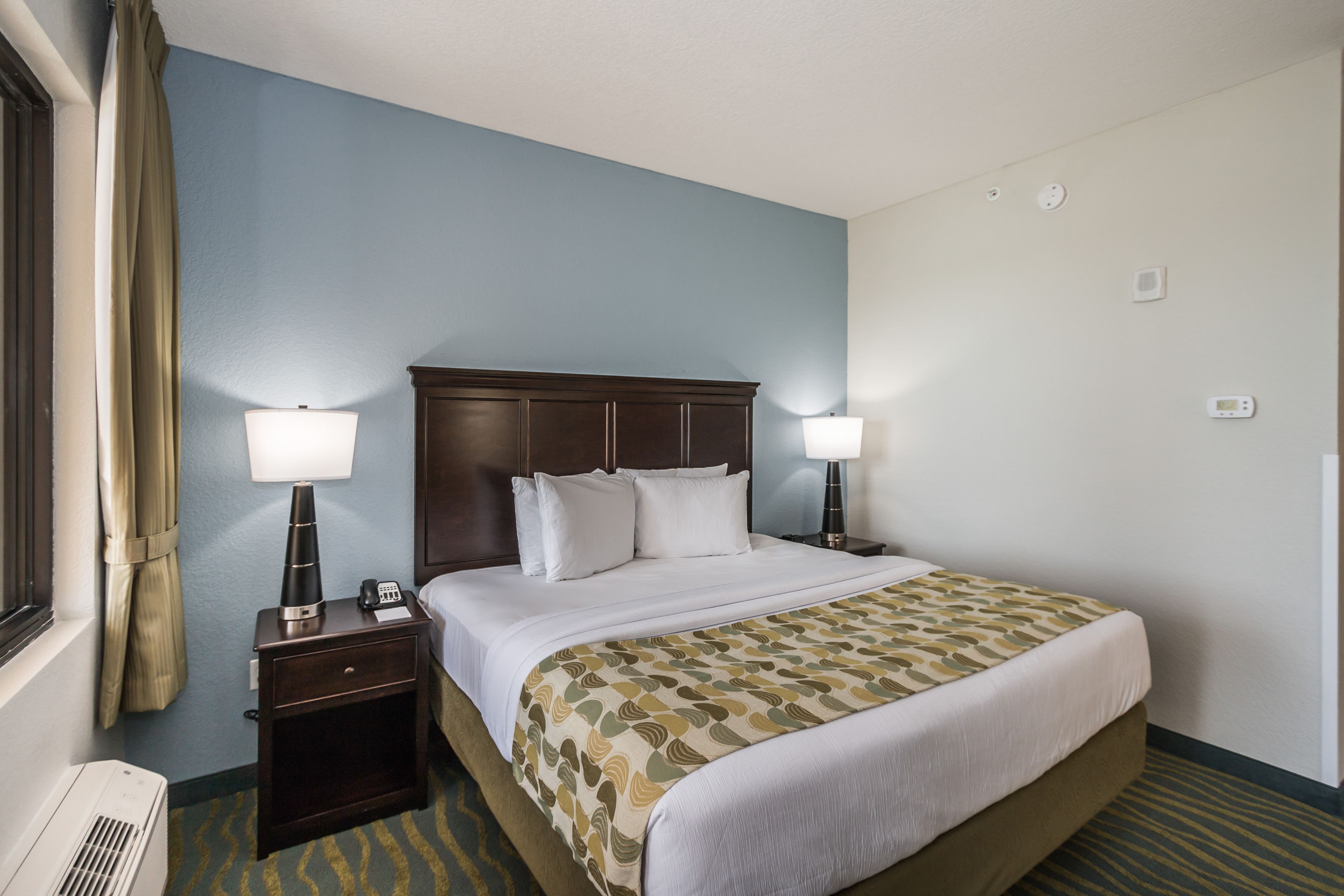 A hotel room with a large bed, two bedside tables, a lamp on each table, a window with curtains, and an air conditioning unit.
