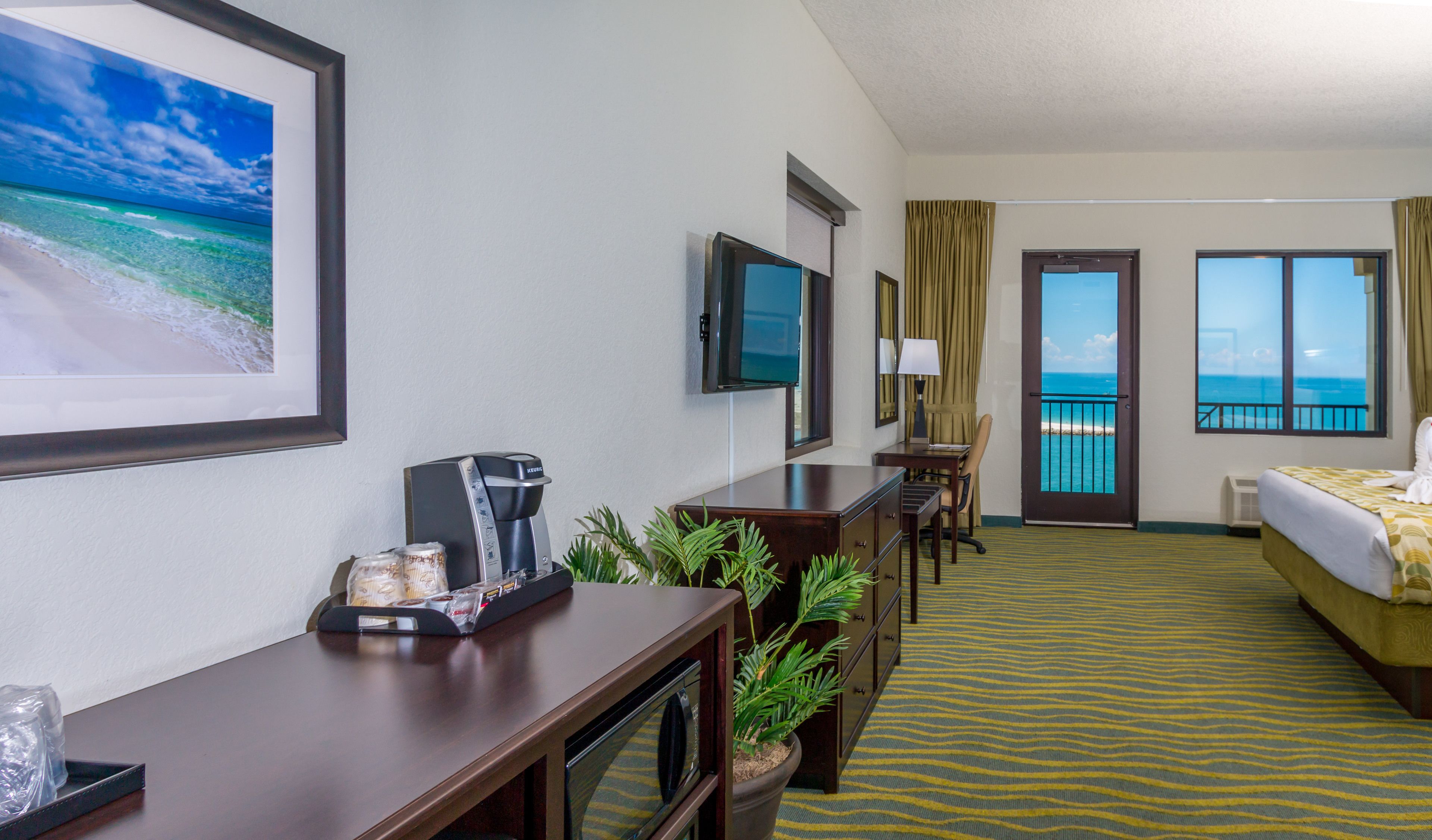 Ein Hotelzimmer mit Bett, Schreibtisch mit Kaffeemaschine, Fernseher, Blumentopf und Meerblick durch eine Tür.