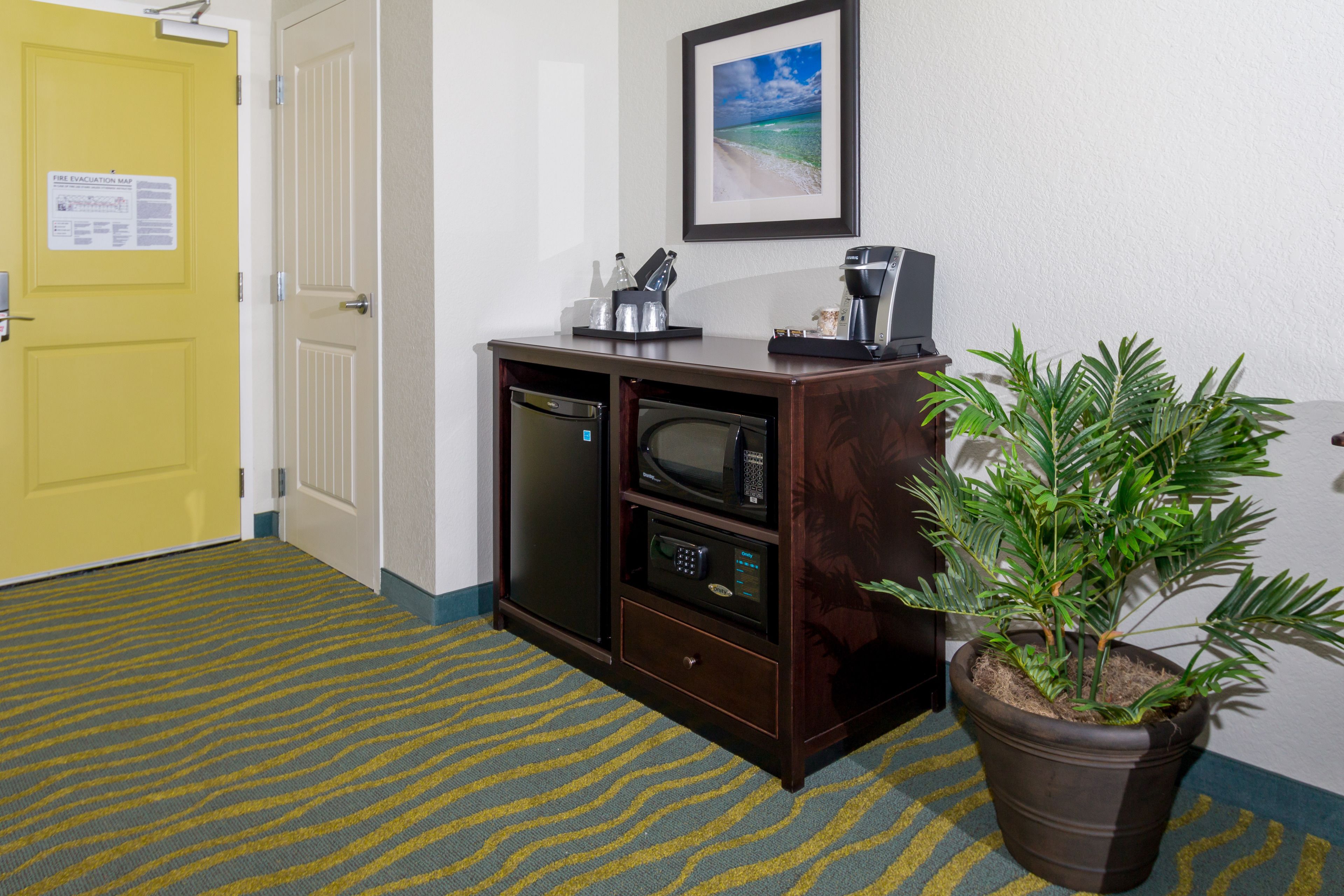 A hotel room with a yellow door, a minibar, a microwave, a coffee maker, and a potted plant.