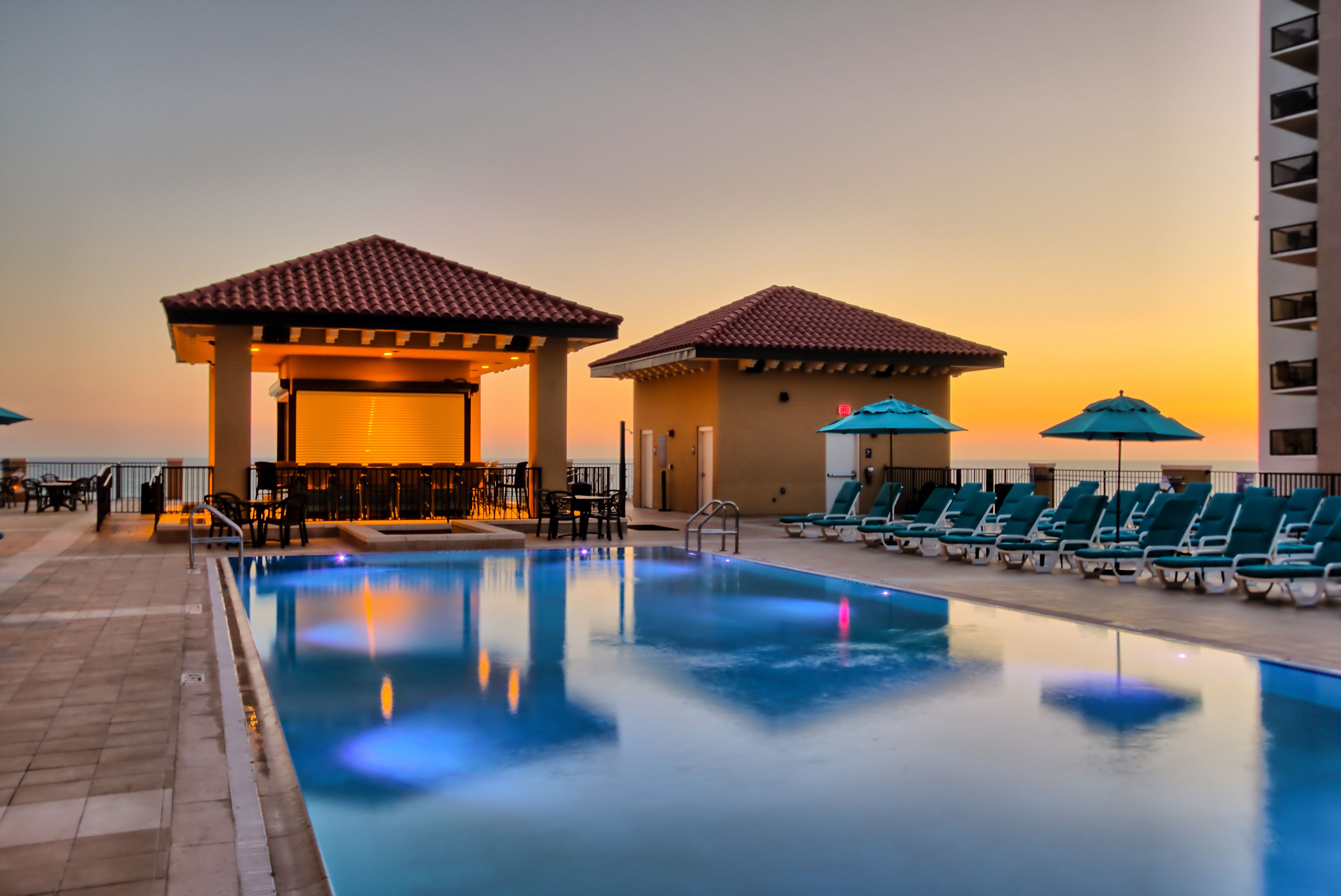 A swimming pool with lounge chairs and umbrellas, a covered seating area, and a view of the sunset.