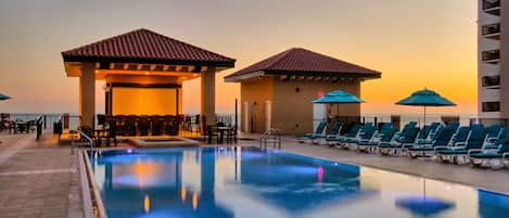 A swimming pool with lounge chairs and umbrellas, a covered seating area, and a view of the sunset.