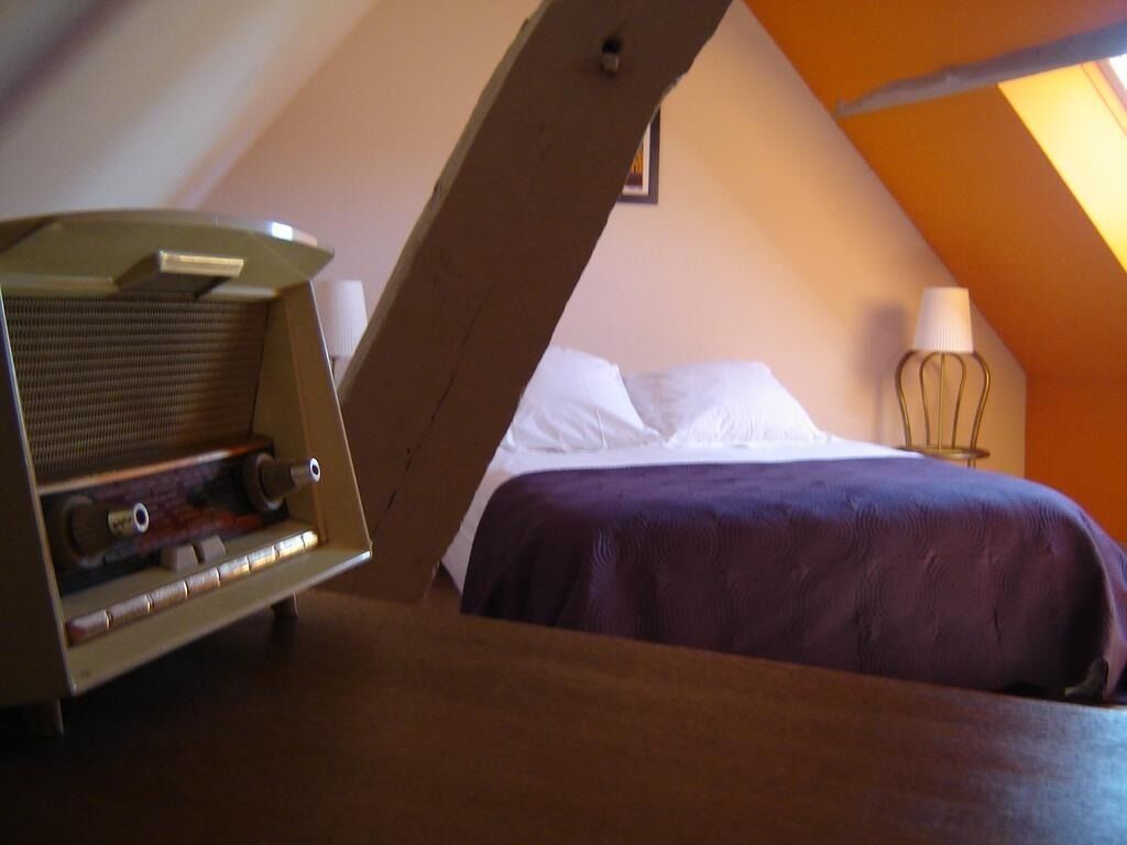 A bedroom with a slanted ceiling, a bed with white and purple bedding, a lamp, and a radio on a wooden table.