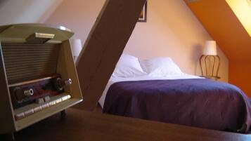 A bedroom with a slanted ceiling, a bed with white and purple bedding, a lamp, and a radio on a wooden table.