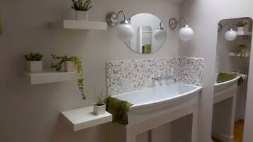 Casa de banho com lavatório branco, espelho e duas luminárias fixadas na parede. Há prateleiras com plantas em vasos sobre o lavatório.