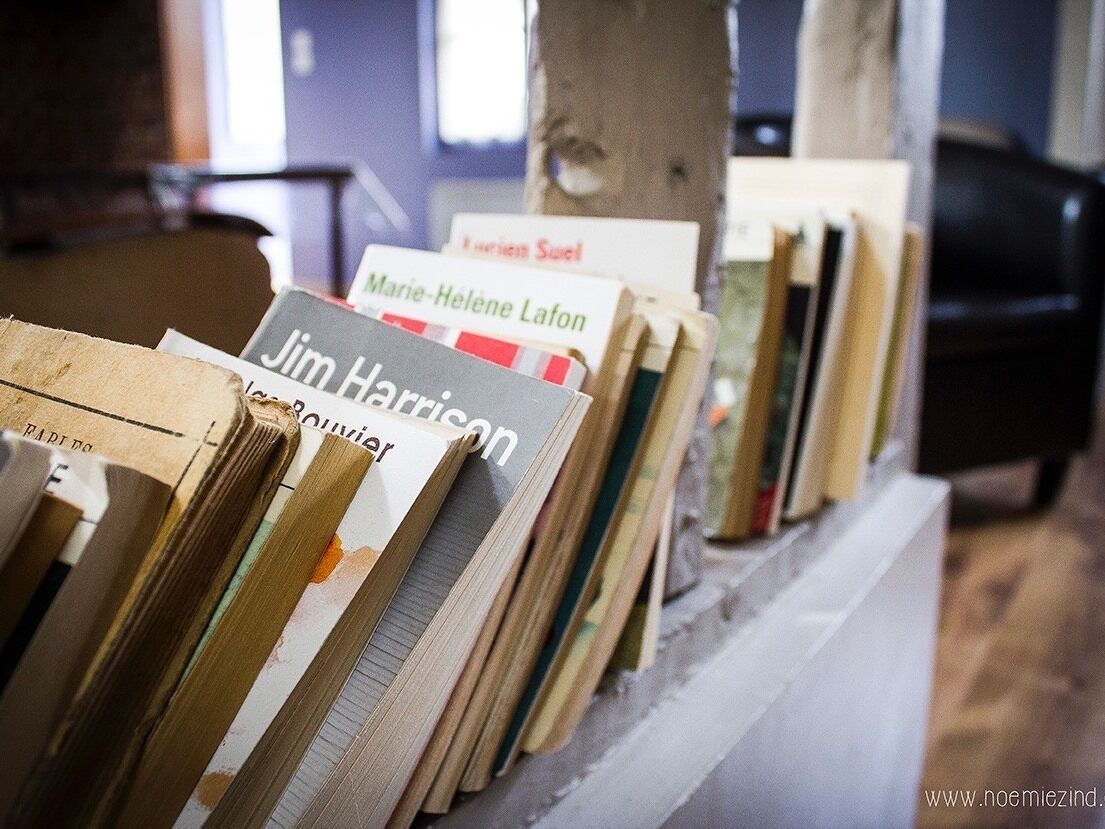 A row of books on a shelf in a room with a concrete pillar and a black chair in the background.